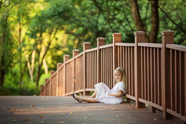 Adorable little girl with long blonde hair sitting on wooden bridge in park sunny summer day. Beautiful child. Happy smiling kid, childhood day, education concept - Photo, Image