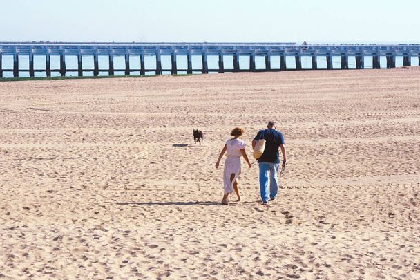 A married couple, a woman in a white dress, and a man walk barefoot along the beach in Ostend. Belgium. Early in the morning by the sea. Wooden bridge against the sky. Traveling together. - Photo, Image