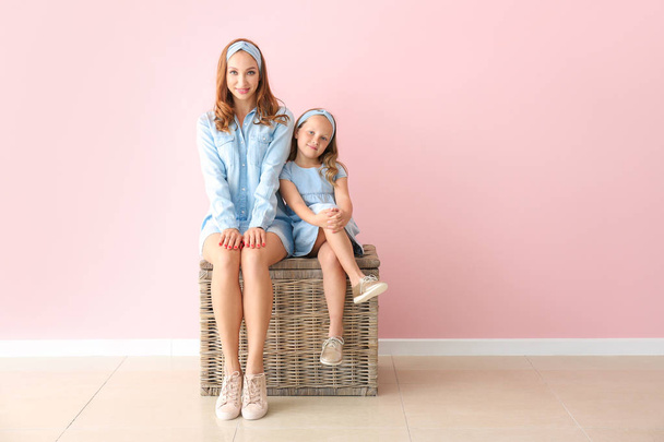 Hermosa mujer y su pequeña hija sentadas en la caja de mimbre cerca de la pared de color
 - Foto, imagen
