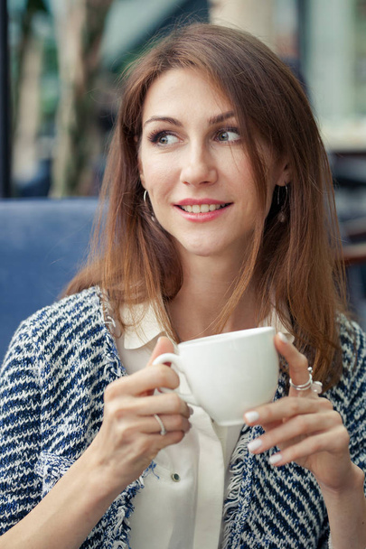 Vertical portrait of beautiful smiling brunette young business woman. The girl holds a white cup in her hands and looks into the distance - Foto, Imagem