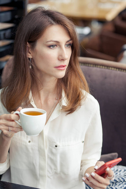 Portrait of a young beautiful brunette business woman in a light shirt. She holds a cup of tea in one hand and a mobile phone in the other. Girl looks thoughtfully into the distance - Foto, Imagen
