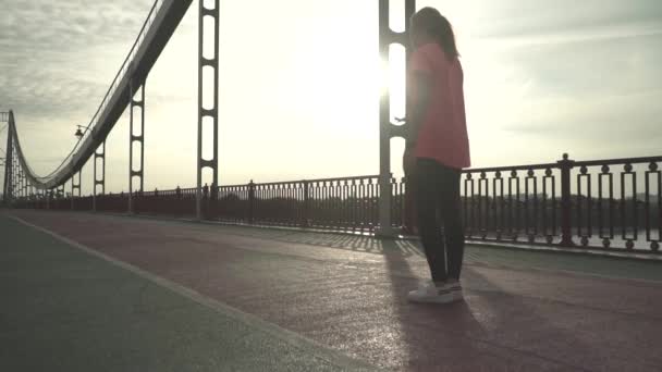 Young athletic girl prepares for morning run on the bridge. The girl is running on the bridge in the morning. The lady spends time outdoor alone. The woman leads a healthy lifestyle. Female moves in slow motion. - Imágenes, Vídeo