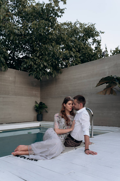 woman in long beautiful dress hugging man while sitting at swimming pool outdoors  - Photo, image