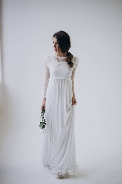 Braut steht auf hellem Hintergrund im weißen Kleid und hält Rosenblume in der Hand  - Foto, Bild