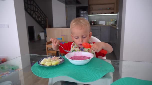 男の子はテーブルの近くの高い椅子に座ってビートルートスープを食べます。ジンバルモーション - 映像、動画