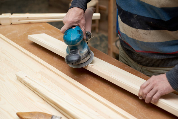 Ένας άνθρωπος μεταχειρίζεται ένα ξύλινο προϊόν με μια μηχανή λείανσης. - Φωτογραφία, εικόνα