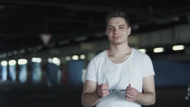Portrait of happy man in white t-shirt wearing sunglasses and standing in the underground parking. Confident man is alone outdoors - Materiał filmowy, wideo