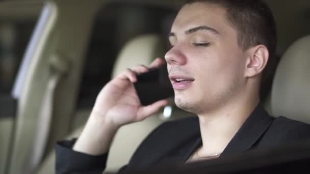 Portrait of young man who is chewing gum and talking by cell phone in the car close up. Leisure of rich confident businessman - Imágenes, Vídeo