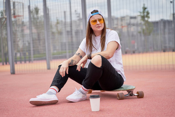 Image of athletic brunette girl in sunglasses sitting on skateboard on playground on summer day - Foto, afbeelding