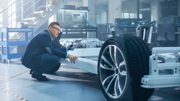 Engineer with Glasses Works on a Tablet Computer Next to an Electric Car Chassis Prototype with Wheels, Batteries and Engine in a High Tech Development Laboratory. - Foto, imagen