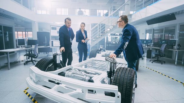 Automotive Design Engineers Talking while Working on Electric Car Chassis Prototype. In Innovation Laboratory Facility Concept Vehicle Frame Includes Wheels, Suspension, Engine and Battery. - 写真・画像