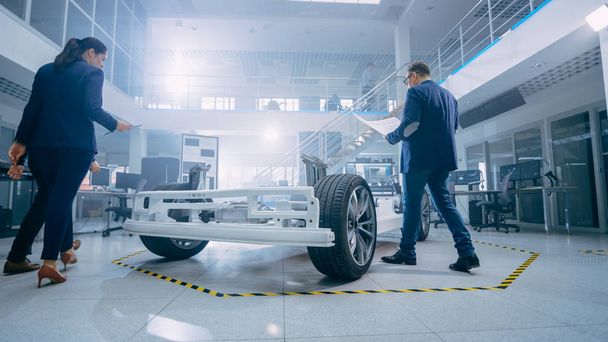 Automotive Design Engineers Talking while Working on Electric Car Chassis Prototype. In Innovation Laboratory Facility Concept Vehicle Frame Includes Wheels, Suspension, Engine and Battery. - Фото, зображення