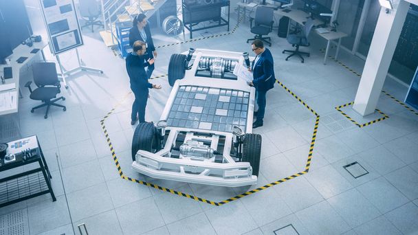 Automotive Design Engineers Talking while Working on Electric Car Chassis Prototype. In Innovation Laboratory Facility Concept Vehicle Frame Includes Wheels, Suspension, Engine and Battery. - Foto, Imagen