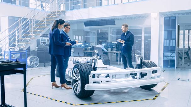 Automotive Design Engineers Looking at Technical Drawings of a Electric Car Chassis Prototype. In Innovation Laboratory Facility Concept Vehicle Frame Includes Wheels, Suspension, Engine and Battery. - Foto, Bild
