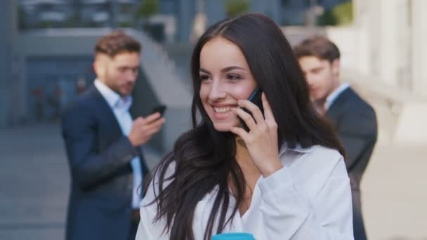 Портрет улыбающейся молодой женщины-предпринимателя, разговаривающей по смартфону возле офисного здания на улицах делового района. Группа занятых бизнесменов на фоне
. - Кадры, видео
