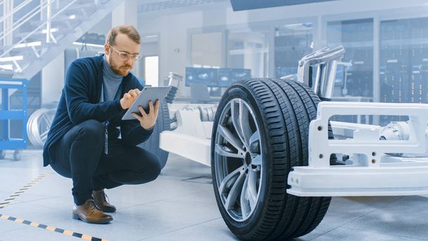 Engineer with Glasses and Beard Works on a Tablet Computer Next to an Electric Car Chassis Prototype with Wheels, Batteries and Engine in a High Tech Development Laboratory. - Foto, immagini
