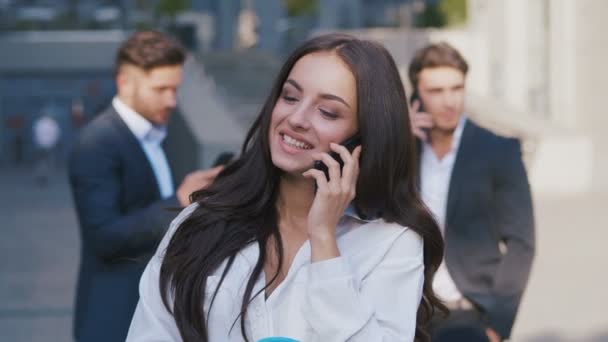 Muotokuva Kaunis nuori nainen Liikenainen puhuu puhelimessa lähellä toimistorakennusta. Tyttö on iloinen ja iloinen. Ryhmä kiireisiä liikemiehiä taustalla
. - Materiaali, video