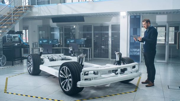 Ingeniero con gafas y barba escanea el prototipo de chasis de coche eléctrico con ruedas, baterías y motor con un software de realidad aumentada en una tableta en un laboratorio de desarrollo de alta tecnología
. - Foto, imagen