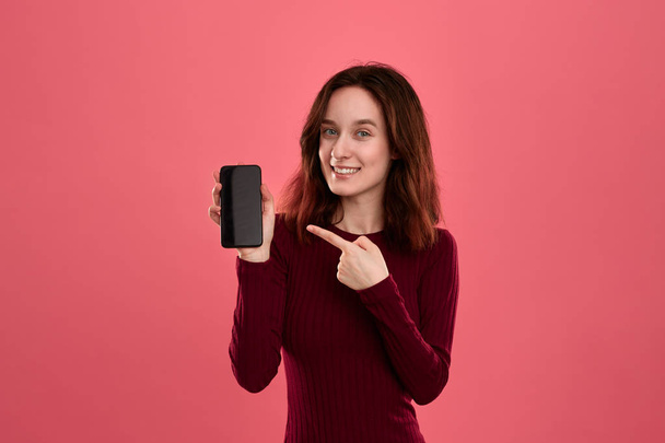 Vzrušená pěkná brunetka ukazující na obrazovku mobilního telefonu prstem, když stojí na tmavě růžovém pozadí a dívá se na kameru. - Fotografie, Obrázek