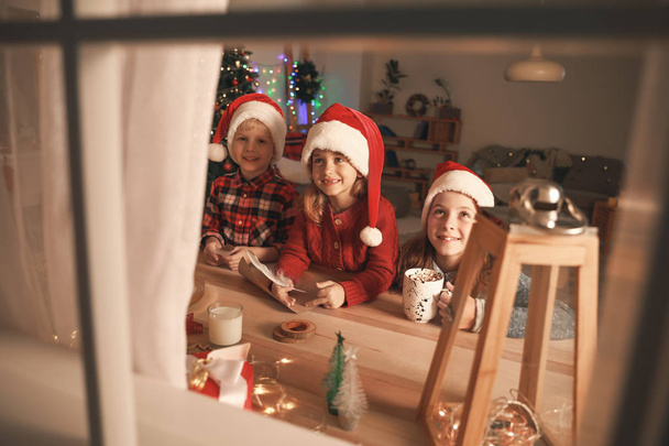 Милые маленькие дети пишут письма Санта Клаусу в канун Рождества дома. Вид через окно
 - Фото, изображение