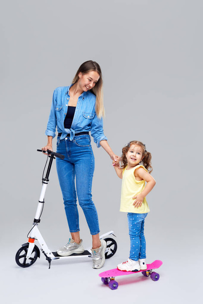 Young woman and little girl standing on scooter and skateboard. Mom teaches daughter to ride, have a fun and joyfully looking in camera over grey background - 写真・画像