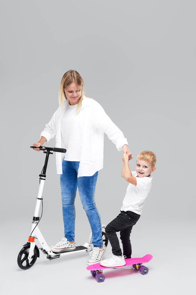 Young woman and child boy standing on scooter and skateboard. Mom teaches son to ride, have a fun and joyfully looking in camera over grey background - Fotografie, Obrázek