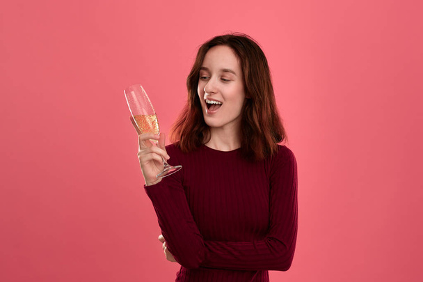 暗いピンクの背景に立ち、カメラに微笑むスパークリングシャンパンのワイングラスを持つ興奮したかわいいブルネットの女の子. - 写真・画像