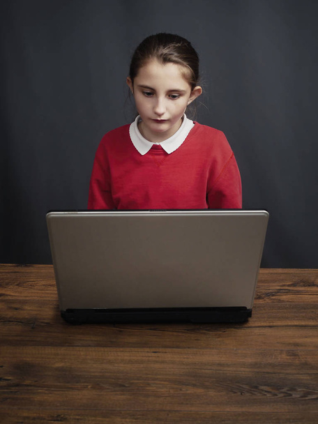 Holka z červené školy dívá se na obrazovku počítače, na tmavém pozadí. Dřevěný stůl. - Fotografie, Obrázek