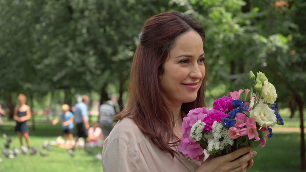 smiling woman walking and smelling flowers in park - Materiał filmowy, wideo