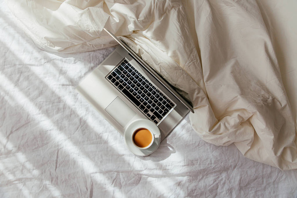 Laptop and cup of coffee on white bed with a  blanket. Work at home concept. Morning light  - 写真・画像