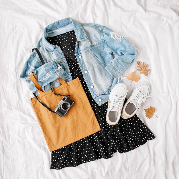 Blue jean jacket and  black dress, sneakers with tote bag and photo camera on white bed. Women's stylish autumn or spring  outfit. Trendy clothes. Flat lay, top view. - Fotografie, Obrázek