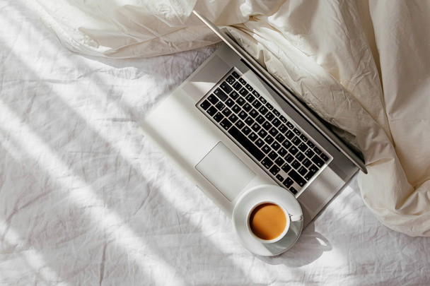 Laptop and cup of coffee on white bed with a  blanket. Work at home concept. Morning light  - Zdjęcie, obraz