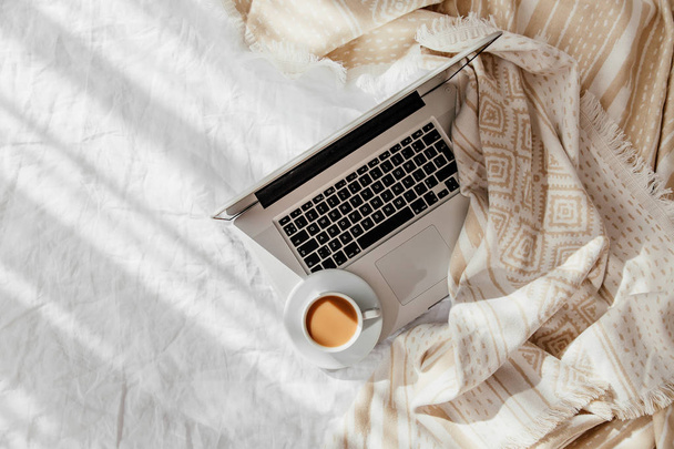 Laptop and cup of coffee on white bed with a beige plaid. Work at home concept. Morning light  - Fotó, kép