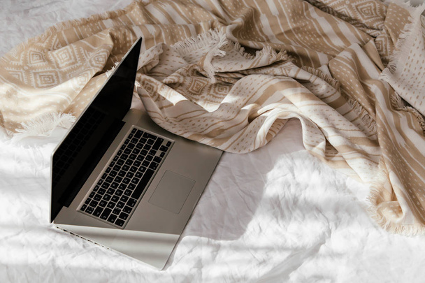 Laptop on white bed with a beige plaid. Work at home concept. Morning light  - 写真・画像