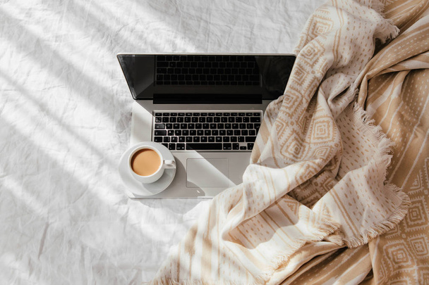 Laptop and cup of coffee on white bed with a beige plaid. Work at home concept. Morning light  - Zdjęcie, obraz