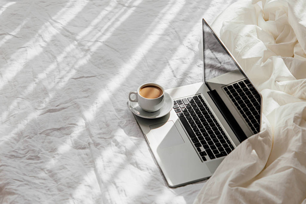 Laptop και φλιτζάνι καφέ σε λευκό κρεβάτι με μια κουβέρτα. Δουλειά στο σπίτι. Πρωινό φως  - Φωτογραφία, εικόνα