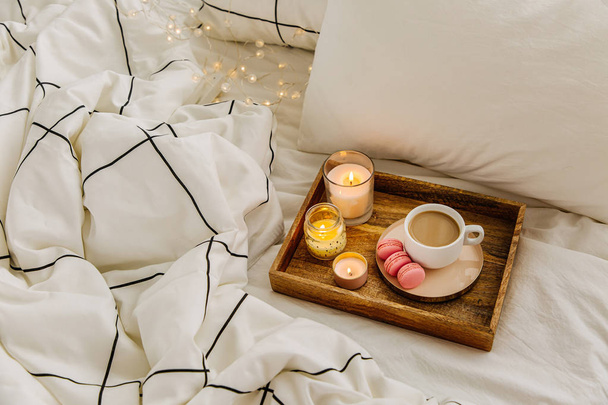 Wooden tray of coffee and candles on bed. White bedding sheets with striped blanket and pillow. Breakfast in bed. Hygge concept. - Foto, Imagen
