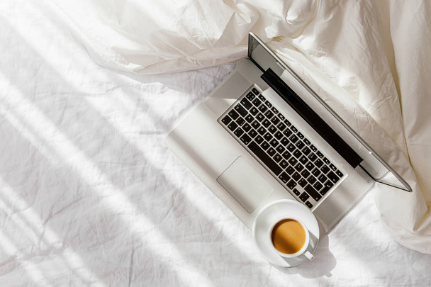 Laptop and cup of coffee on white bed with a  blanket. Work at home concept. Morning light  - Fotografie, Obrázek