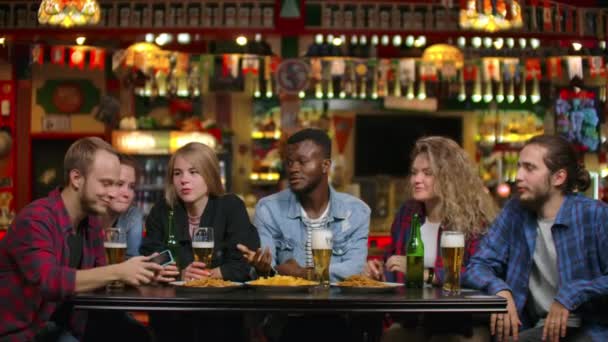 Duża grupa przyjaciół w barze siedząc przy stole śmieje się i rozmawiając pijąc piwo - Materiał filmowy, wideo