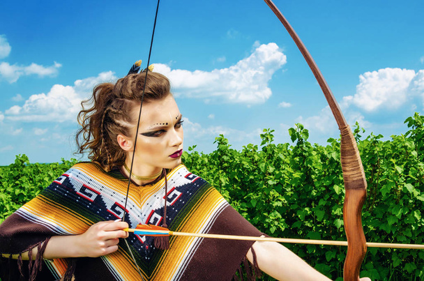 Amazon girl aggressive makeup, in a poncho, cosplay in the field, holds a bow and arrow. Paganism, feminism, independence, tribe - Photo, Image