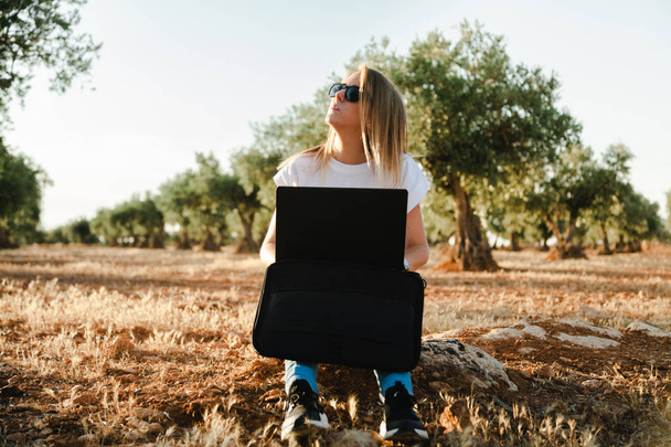 Mujer joven con una funda para portátil sentada en el campo de olivos
 - Foto, Imagen