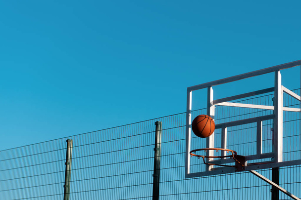 Basketball ball is hitting the target. Ball is flying to the basketball hoop against a blue sky with a metal fence. Success concept photo. Copy space. Great teamwork. - Фото, изображение
