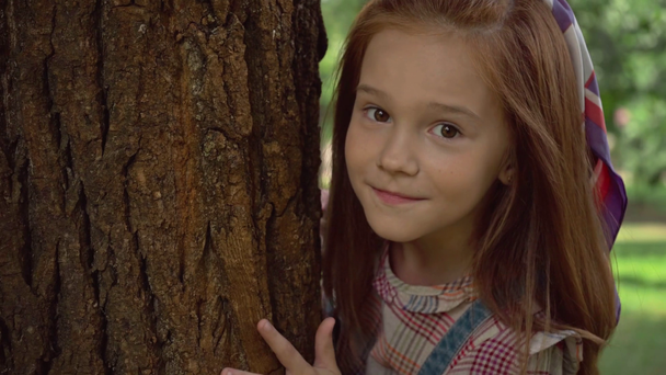 cute redhead child hiding behind tree trunk and showing tongue - Imágenes, Vídeo