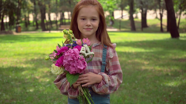 ευτυχισμένο κοκκινομάλλα παιδί μυρωδιά λουλούδια σε πράσινο πάρκο - Πλάνα, βίντεο