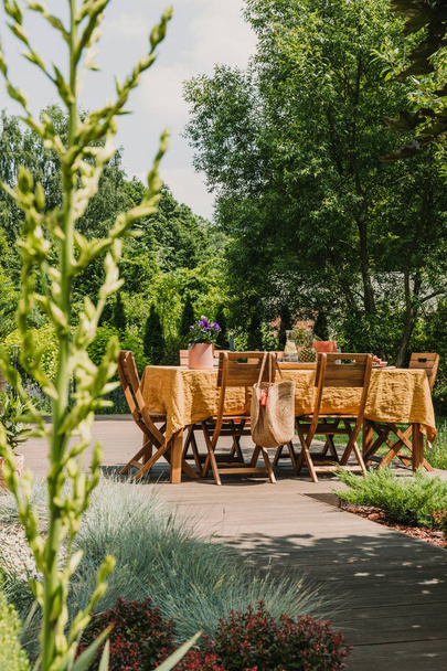 Pystysuora näkymä ruokapöytä peitetty oranssi pöytäliina seisoo puinen terassi vihreässä puutarhassa
 - Valokuva, kuva