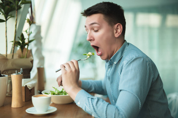 Молодой красивый мужчина кладет салат в рот и пьет кофе на обед во время перерыва в кафе рядом с офисом выглядит голодным
 - Фото, изображение