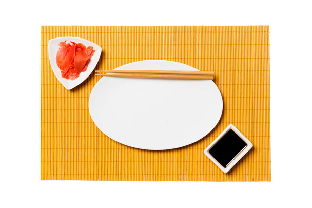 Üres ovális fehér lemez pálcika a sushi és a szójaszósz, gyömbéres sárga bambusz szőnyeg háttérben. Felülnézet másolási területtel a tervezéshez - Fotó, kép