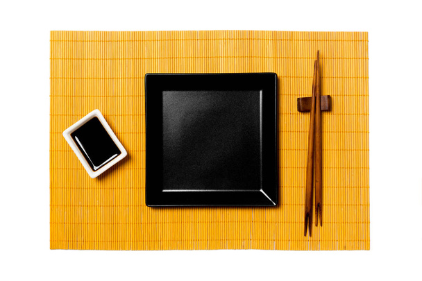 Üres fekete négyzet alakú tányér pálcika a sushi és a szójaszósz sárga bambusz szőnyeg háttér. Felülnézet másolási területtel a tervezéshez - Fotó, kép