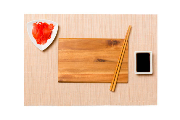 Lege rechthoekige bruine houten plaat met stokjes voor sushi, gember en sojasaus op bruin Sushi mat achtergrond. Bovenaanzicht met Kopieer ruimte voor uw ontwerp - Foto, afbeelding
