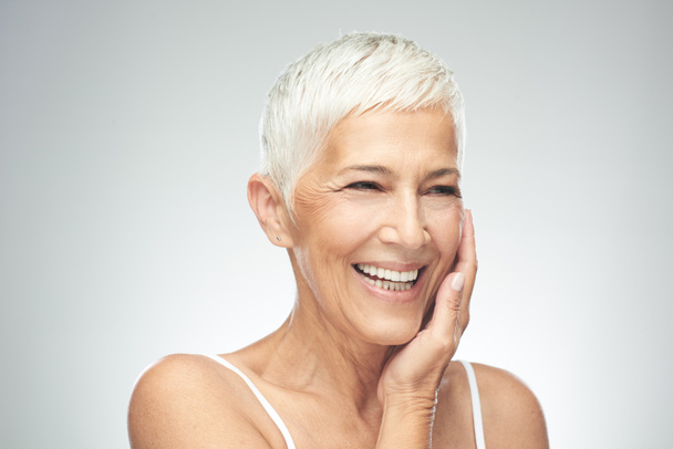 Mooie lachende Senior vrouw met korte grijze haren poseren in de voorkant van de grijze achtergrond. Beauty fotografie. - Foto, afbeelding